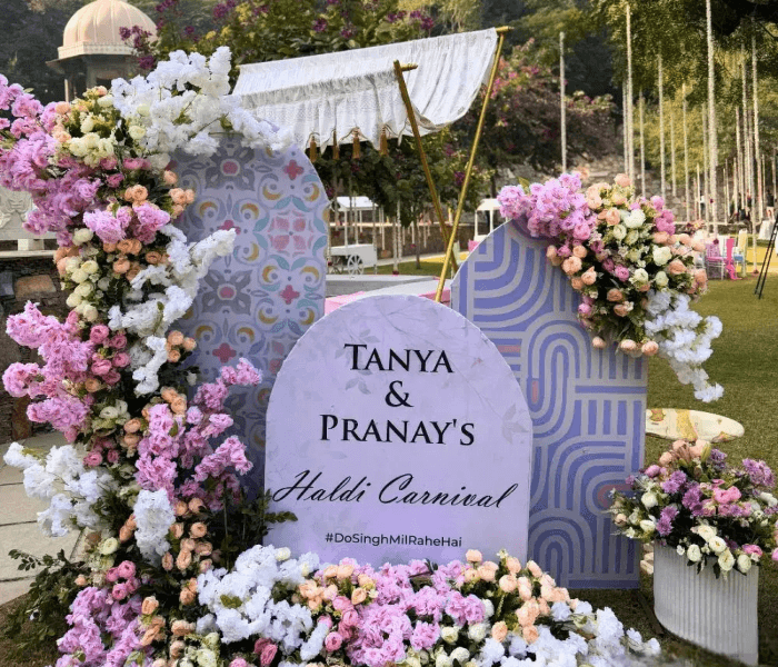 Modern Fusion Carnival - Happy Wedding App Haldi carnival welcome sign reading Tanya & Pranay's Haldi Carnival, surrounded by pink and white floral arches.