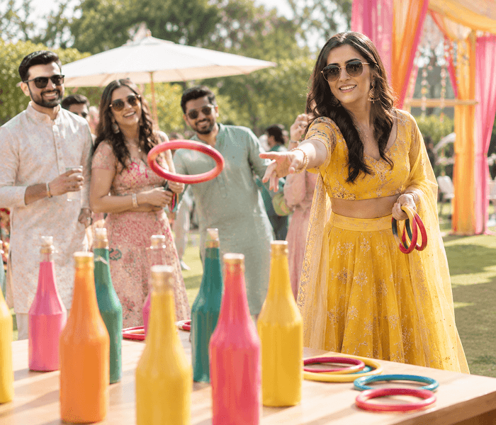 Ring Toss - Happy Wedding App A smiling woman in a yellow outfit tossing a ring toward colorful bottles arranged on a table.